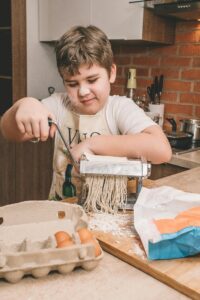 A young boy joyfully prepares homemade pasta using a pasta machine in a cozy kitchen setting.