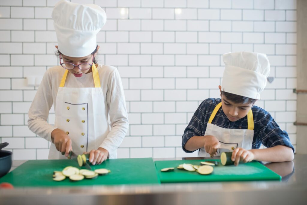 Two kids learning to cook by slicing vegetables indoors wearing chef hats and aprons.